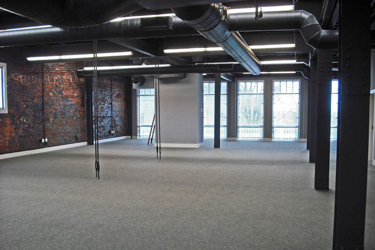 Renovated Wick's Building 3rd Floor shows an open floor plan with an exposed brick wall, black ventilation system on the ceiling, and a wall of windows along the back.