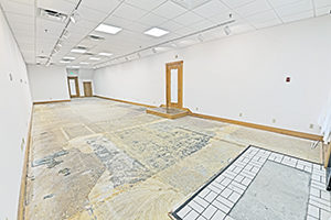 Uptown Plaza, Suite 115, view 2, front to back, white walls, bright lighting, elongated space, unfinished floor. Space offers a second entrance off of the main lobby. Second door has a small platform.